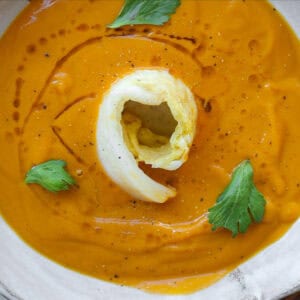 A bowl of gochujang and roasted carrot soup, garnished with a swirl of cream and fresh celery leaves. The soup is in a white bowl placed on a patterned surface with hints of garlic visible nearby. square